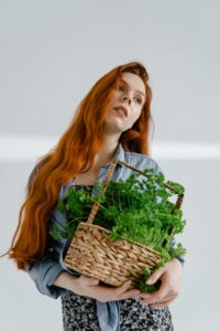 Elegant young woman with long red hair holding a basket of fresh greens in a bright, serene setting.